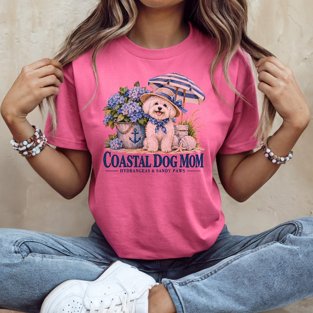 A person wearing a pink t-shirt with a graphic of a dog, hydrangeas, umbrella, and text reading Coastal Dog Mom.