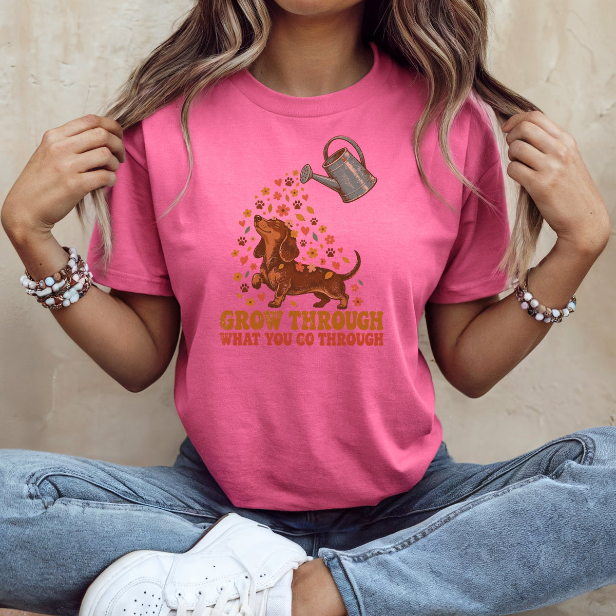 A woman wearing a pink t-shirt printed with a dachshund being watered by a can and the text GROW THROUGH WHAT YOU GO THROUGH.