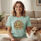 A smiling woman wears a mint green t-shirt with a golden retriever graphic, sitting beside a small terrier.
