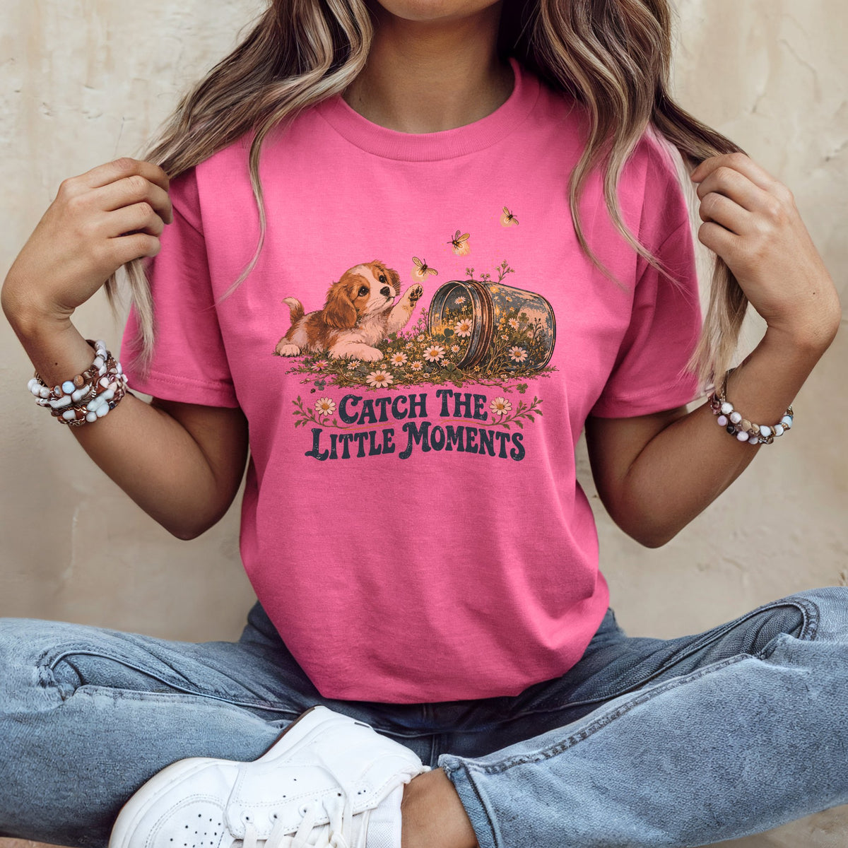 A person wearing a bright pink t-shirt with a graphic of a puppy, flowers, bees, and the text "Catch the Little Moments."