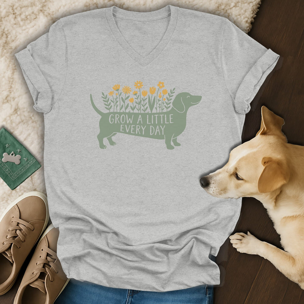 Gray V-neck t-shirt featuring a green dachshund graphic with flowers and text, laid flat with accessories and a small dog.