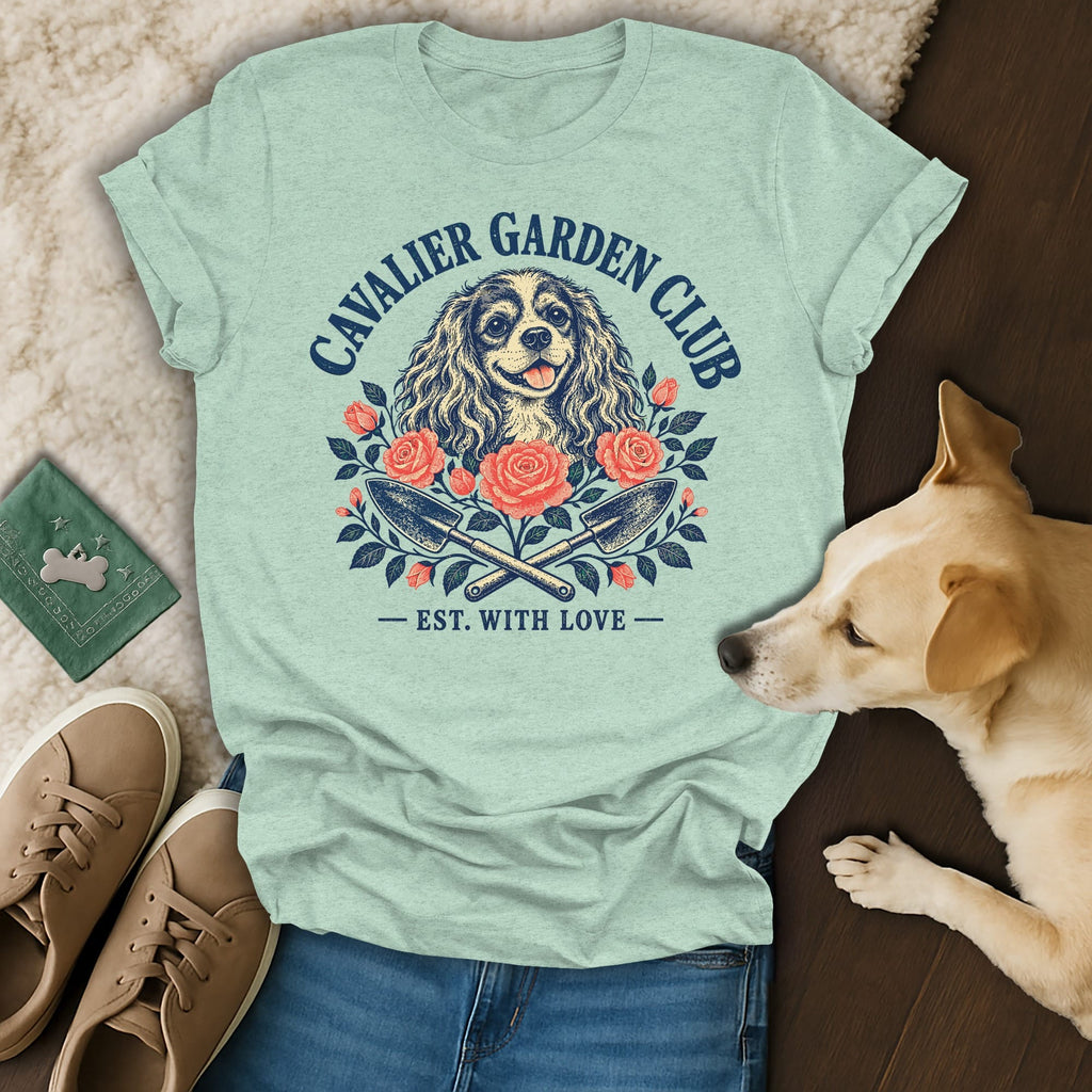 A light green t-shirt with a Cavalier Garden Club graphic featuring a dog, roses, and crossed gardening trowels.