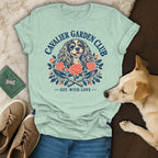 A light green t-shirt with a Cavalier Garden Club graphic featuring a dog, roses, and crossed gardening trowels.