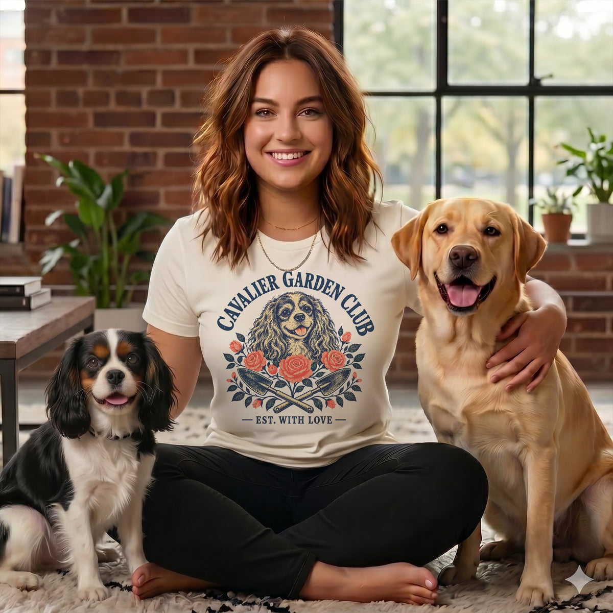 Smiling woman wearing a t-shirt with a dog graphic sits flanked by a Cavalier King Charles Spaniel and a Labrador Retriever.