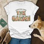 A white t-shirt featuring floral lettering that reads Greetings From The Garden surrounding a corgi illustration, laid out with shoes, jeans, and a real dog.
