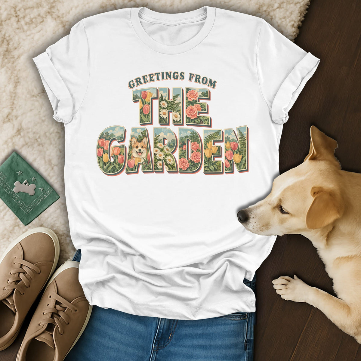 A white t-shirt featuring floral lettering that reads Greetings From The Garden surrounding a corgi illustration, laid out with shoes, jeans, and a real dog.