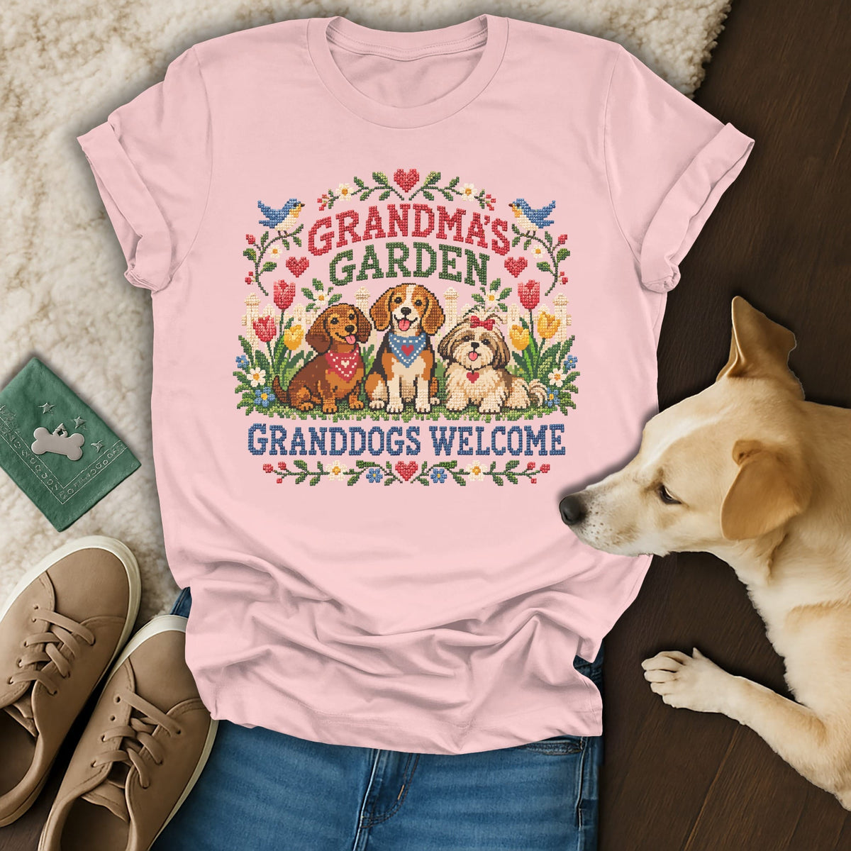A pink t-shirt featuring a cross-stitch graphic of three dogs, flowers, and Grandma's Garden Granddogs Welcome text.