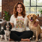 A smiling woman sits cross-legged on a rug between a Cavalier King Charles Spaniel and a Labrador Retriever, wearing a t-shirt with a graphic of a dog surrounded by flowers and the text GRAY HAIR DON'T CARE.