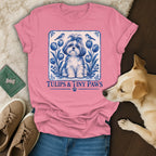 A pink t-shirt featuring a blue-and-white graphic of a dog, tulips, and birds, with the text Tulips & Tiny Paws.