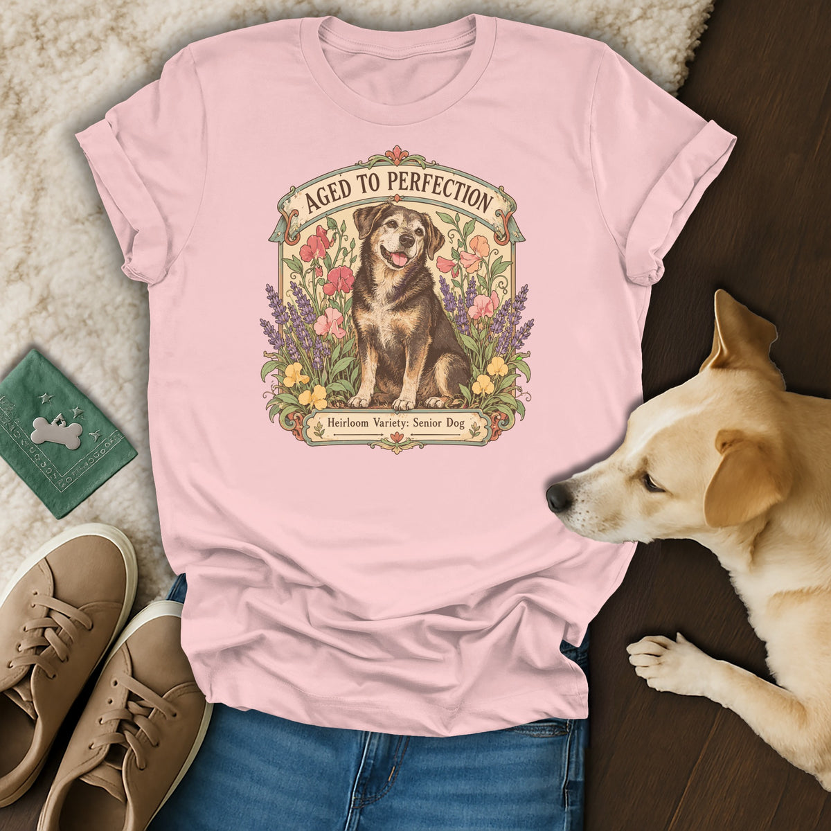 Pink t-shirt with a vintage dog graphic reading "Aged to Perfection" surrounded by flowers, placed among accessories and a real dog.