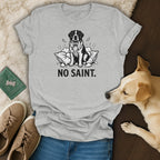 Grey t-shirt with black line art of a dog sitting on pillows, text "NO SAINT."