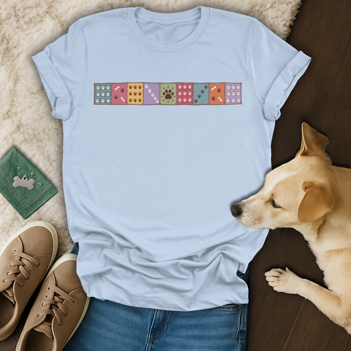 Light blue t-shirt with a colorful paw print patchwork graphic, laid out with blue jeans, brown sneakers, and a small dog.