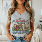 A woman wearing a gray V-neck t-shirt featuring a cross-stitch design of three dogs and flowers with the text "GRANDMA'S GARDEN GRANDDOGS WELCOME".