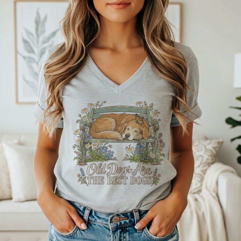 A woman wearing a gray V-neck t-shirt featuring a graphic of a sleeping dog on a bench surrounded by flowers and the text Old Dogs Are The Best Dogs.