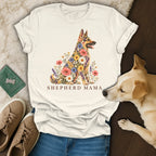 A cream-colored t-shirt featuring a floral German Shepherd illustration and the text SHEPHERD MAMA, laid out on a dark surface with casual shoes and a dog resting nearby.