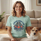 A smiling woman wearing a mint-green t-shirt featuring a Cavalier King Charles Spaniel graphic, sitting indoors beside a terrier.