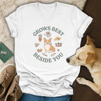 A white V-neck t-shirt featuring a corgi and garden graphic with the text Grows Best Beside You.