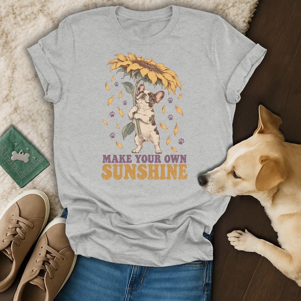 A light grey t-shirt featuring a graphic of a French Bulldog holding a sunflower, with text reading MAKE YOUR OWN SUNSHINE.