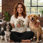 A smiling woman wearing a graphic t-shirt sits cross-legged between two dogs indoors.