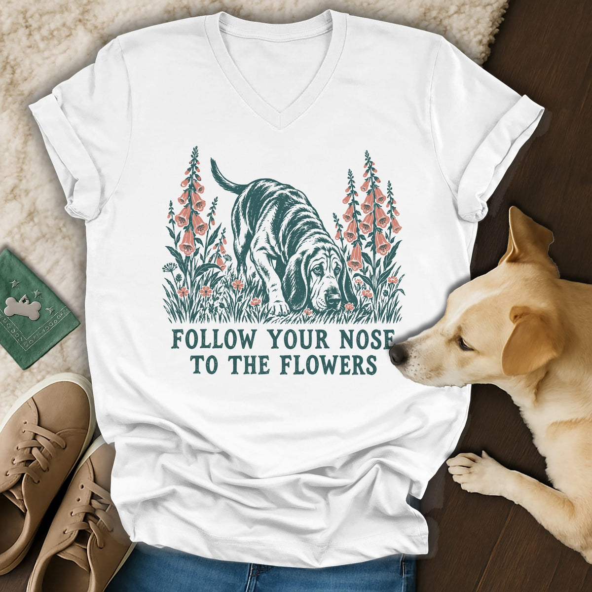 A white V-neck t-shirt featuring a vintage-style graphic of a basshound sniffing flowers with the text "FOLLOW YOUR NOSE TO THE FLOWERS".