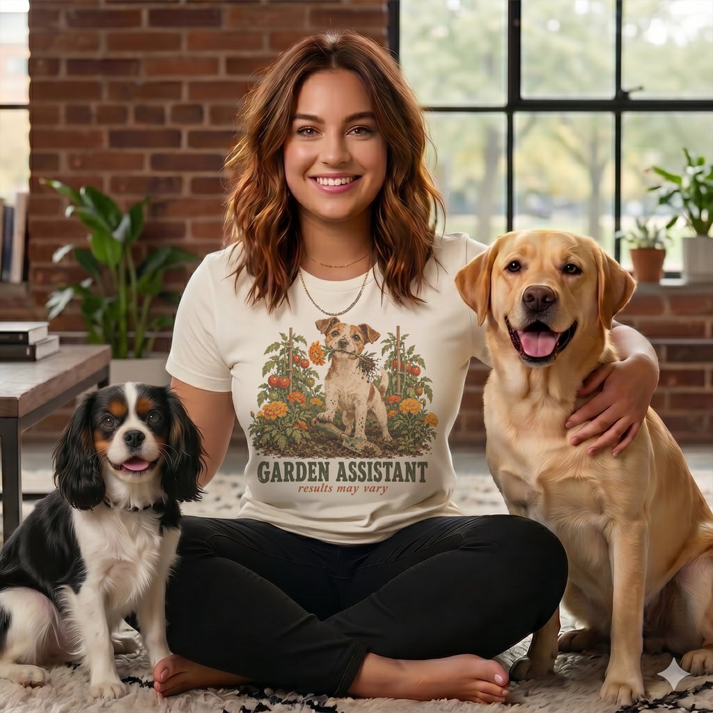 A smiling woman wearing a garden assistant graphic t-shirt sits cross-legged between two dogs.