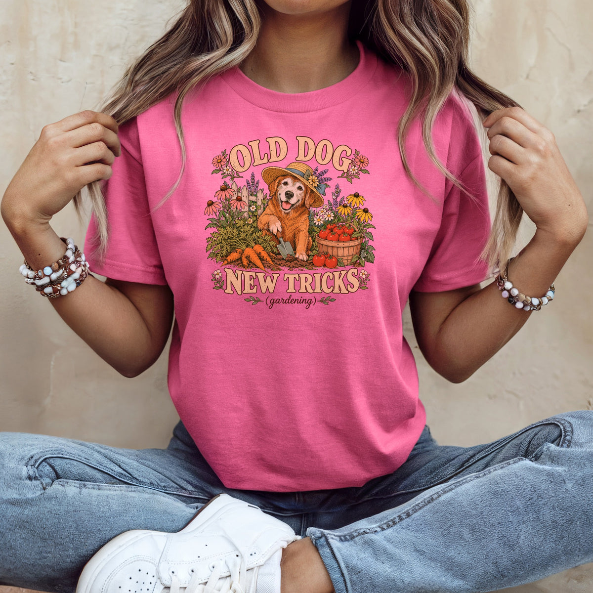A woman sitting down wearing a pink t-shirt with a graphic of a gardening dog and the text Old Dog New Tricks.
