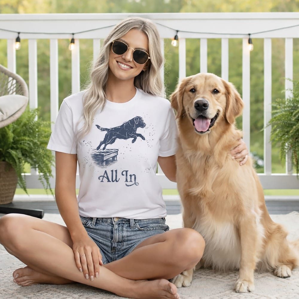 Woman in white t-shirt with "All In" text and jumping dog graphic sits with Golden Retriever.
