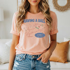 Peach t-shirt with cartoon dog playing with balls, reading "Having a ball is kind of my thing."