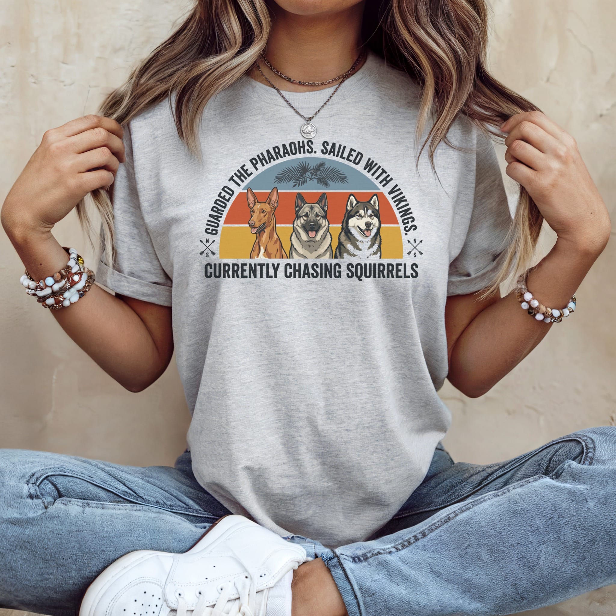 Grey t-shirt with a graphic of three dogs and text "Guarded the pharaohs, sailed with Vikings. Currently chasing squirrels."