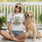 A smiling blonde woman wearing sunglasses and a white graphic t-shirt sits cross-legged next to a golden retriever on a porch.