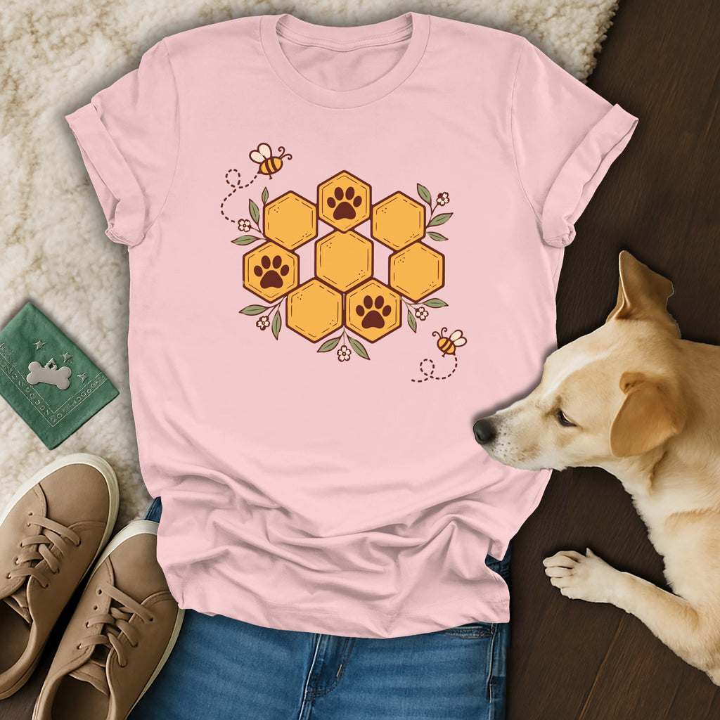 A pink t-shirt featuring a honeycomb graphic with paw prints, surrounded by bees and leaves, lies on a fluffy rug next to a small dog, sneakers, and a green passport case.