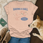 Light peach t-shirt with cartoon dog playing with balls and text "HAVING A BALL is kind of my thing."