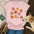 A pink t-shirt featuring a graphic of autumn leaves shaped like paw prints, surrounded by a dog, shoes, and a green tag.