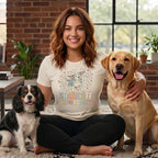 A smiling woman wearing a graphic t-shirt sits cross-legged between two dogs on the floor.