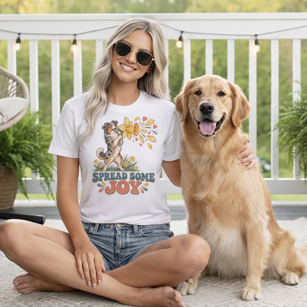A smiling woman wearing a white graphic t-shirt sits cross-legged beside a golden retriever.