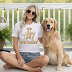Woman wearing white t-shirt with "STAY GOLDEN" and a graphic, sitting with a golden retriever.