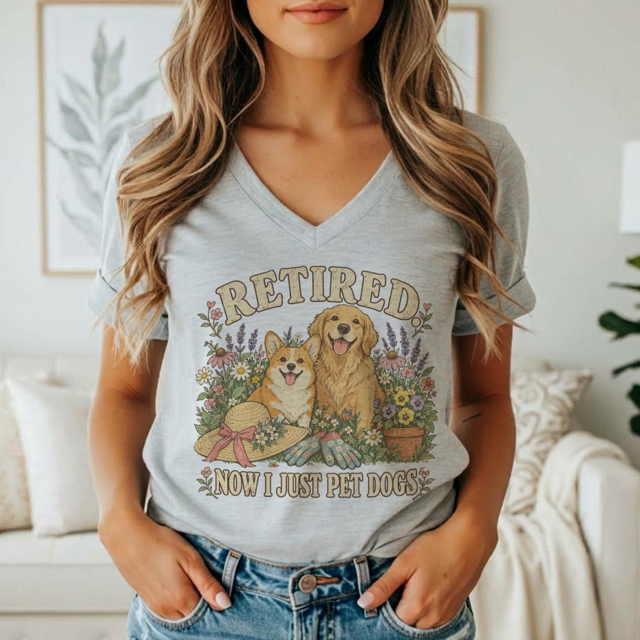 A woman wearing a heather gray V-neck t-shirt with a graphic of dogs and flowers that reads Retired Now I Just Pet Dogs.