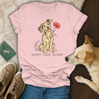 A pink t-shirt featuring an illustration of a dog holding a red flower and the text "ADOPT. LOVE. BLOOM."