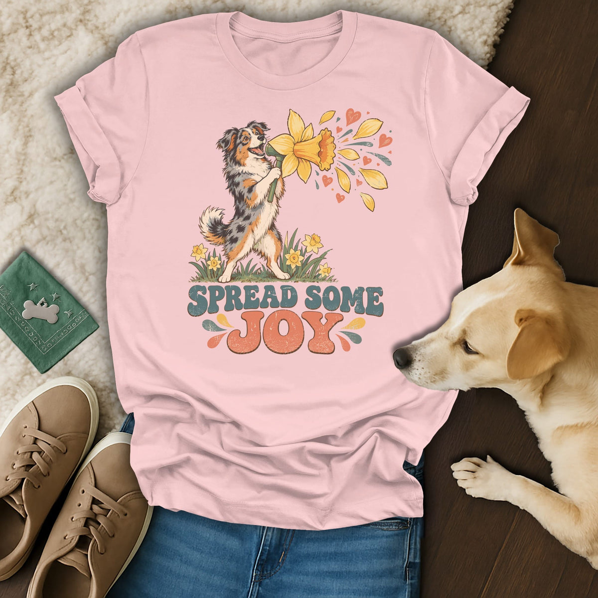 A pink t-shirt featuring an Australian Shepherd graphic and the text "Spread Some Joy," surrounded by shoes, a green wallet, and a dog lying nearby.