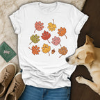 A white t-shirt featuring an autumn leaf and paw print graphic, surrounded by casual accessories and a dog.