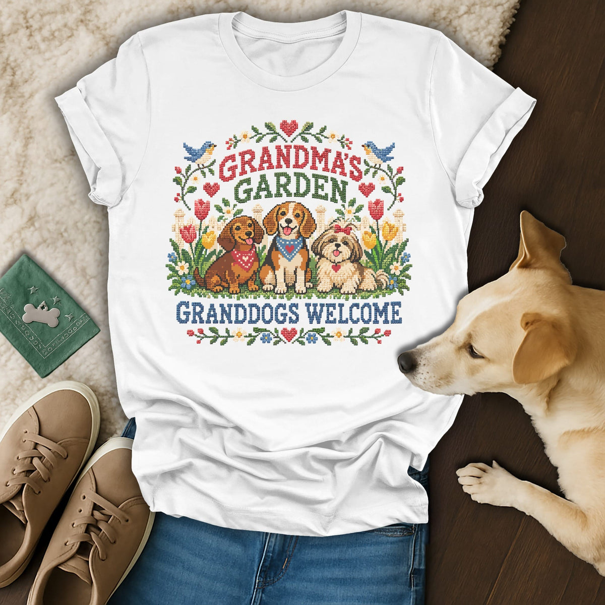A white crewneck t-shirt featuring a cross-stitch graphic of three dogs surrounded by flowers, with the text Grandma's Garden Granddogs Welcome.