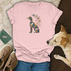 A pink t-shirt featuring a stained-glass style dog graphic with floral accents, laid out with blue jeans, tan shoes, and a real dog.