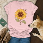 A light pink t-shirt with a sunflower and paw print graphic, arranged flat with a dog, sneakers, and accessories.