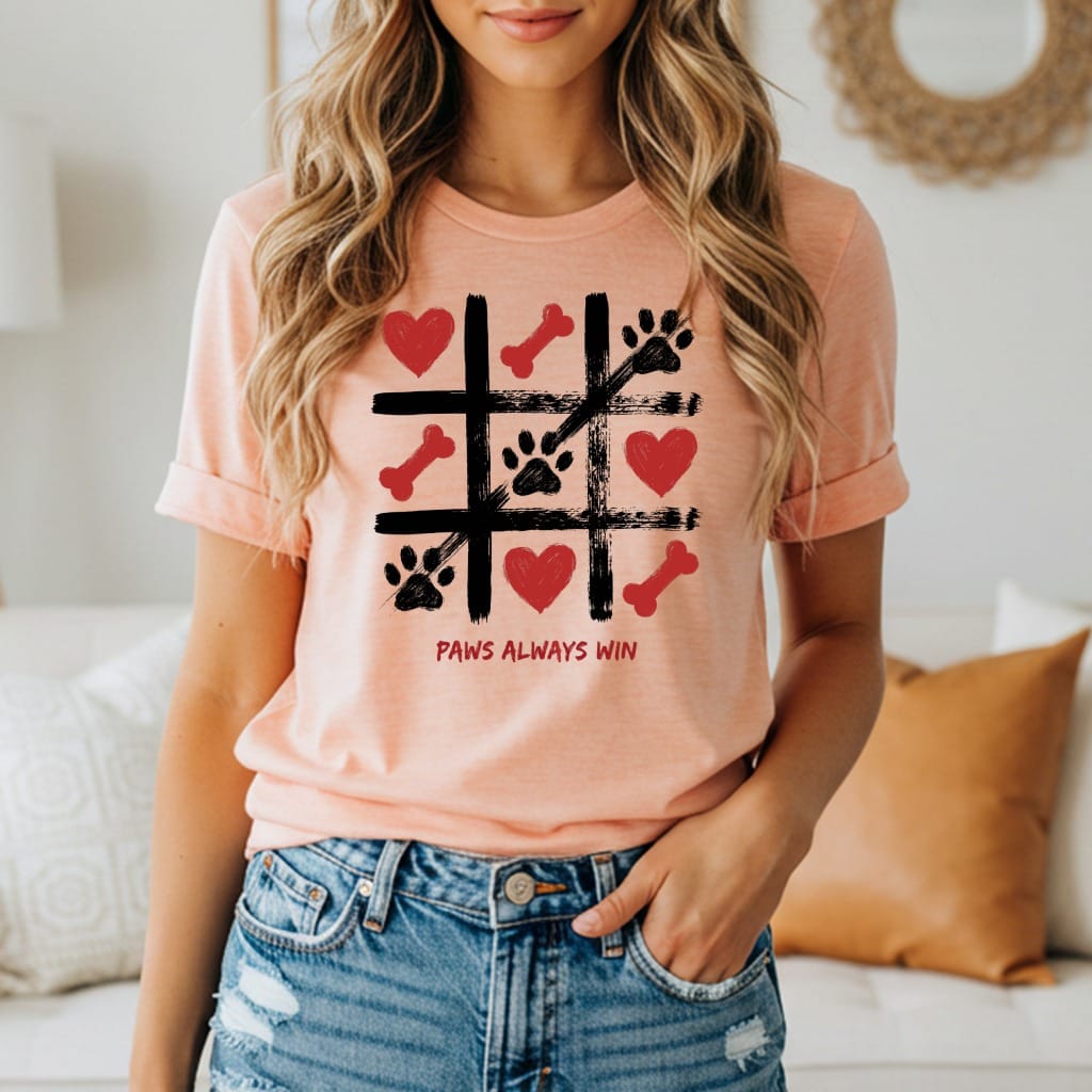 Peach t-shirt with a tic-tac-toe graphic featuring paw prints, hearts, and bones, with text "Paws Always Win".