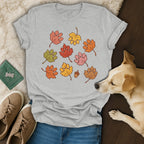 Gray crewneck t-shirt with autumn leaf paw print graphics, placed on a wooden floor alongside shoes and a dog.